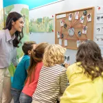 Une enseignante et un groupe d’enfants observent un panneau d’enquête affiché au mur, avec des cartes, photos et fils rouges reliant différents éléments. À côté, des fiches illustrées sont affichées, présentant des informations sur la nature. L’ensemble évoque un travail d’investigation coopératif autour de l’environnement.