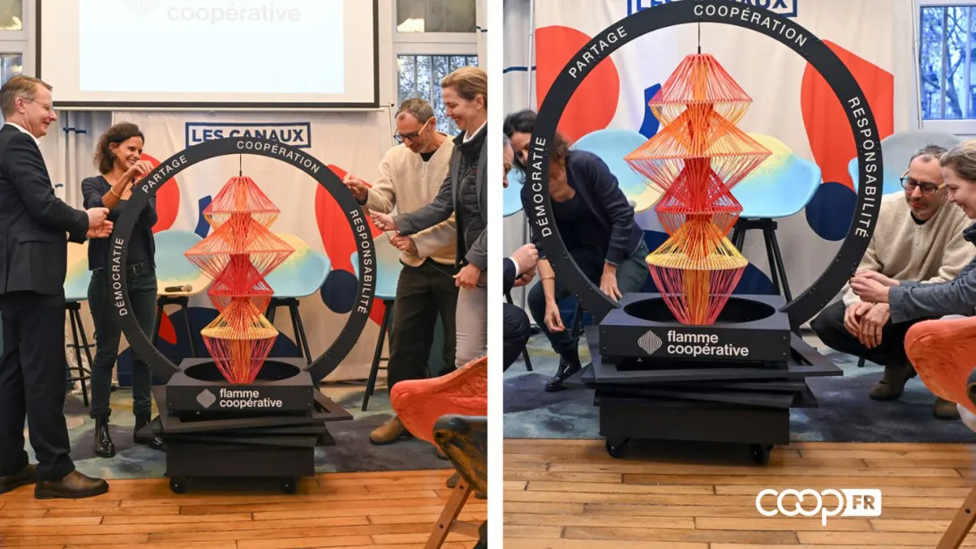 Événement de présentation de la ‘Flamme Coopérative’ aux Canaux. Plusieurs personnes sont réunies autour d’une installation artistique composée de fils tendus formant une structure géométrique, encadrée par un anneau portant les mots ‘Démocratie’, ‘Partage’, ‘Coopération’ et ‘Responsabilité’. Les participants échangent et posent pour des photos officielles.