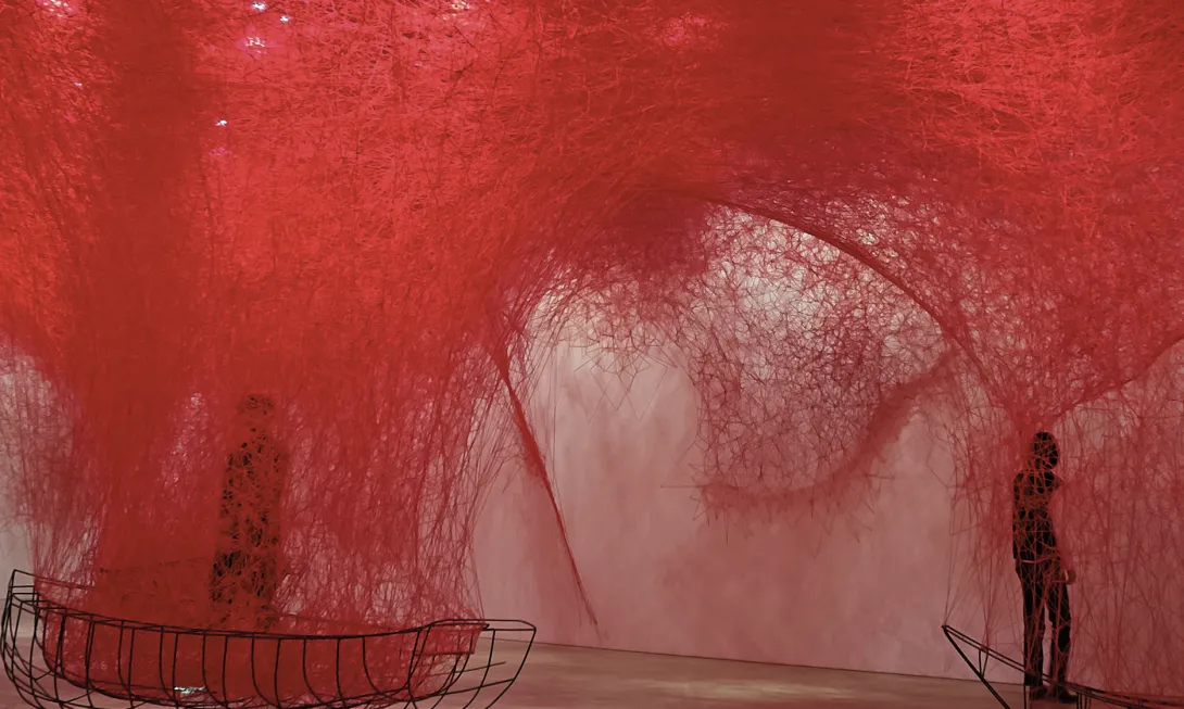 Installation artistique immersive composée d’un enchevêtrement dense de fils rouges suspendus, créant une atmosphère onirique et enveloppante. Deux silhouettes humaines se tiennent à l’intérieur de l’œuvre, accentuant l’effet de profondeur et de mystère. Des structures métalliques en forme de barques sont disposées au sol, renforçant l’idée d’un voyage ou d’une traversée symbolique.